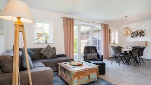 A living room with a sofa, a coffee table, a lamp, a dining area with chairs and a table, and a view through a glass door. - Fischhuus - Beautiful vacation home near the water (Krummhörn)