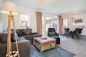 A living room with a sofa, a coffee table, a lamp, a dining area with chairs and a table, and a view through a glass door. - Fischhuus - Beautiful vacation home near the water (Krummhörn)