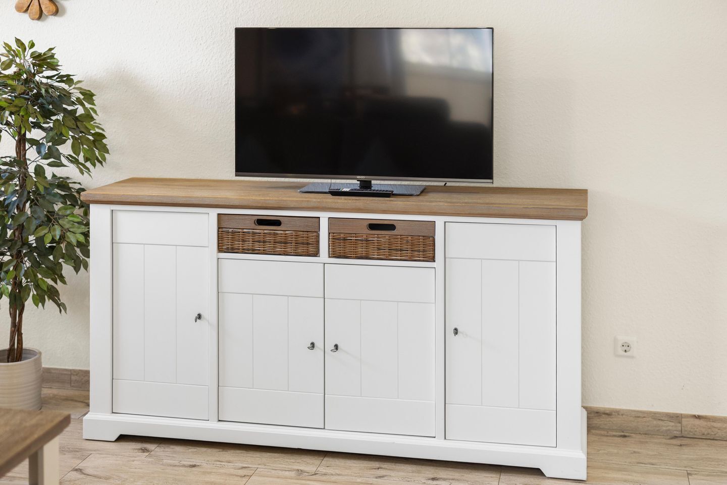 A white cabinet with a wooden top, a flat-screen TV mounted above it, and two wicker baskets on the cabinet doors.