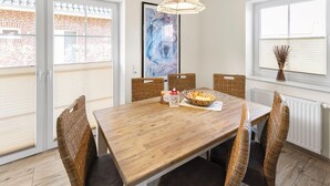 A dining area with a wooden table and wicker chairs, a painting on the wall, and a window with blinds. - Udo - Schöne Ferienwohnung in Zentraler Lage (Krummhörn)