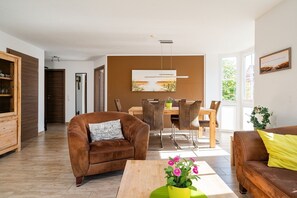 A modern living room with a brown leather sofa, a wooden coffee table with a vase of pink flowers, and a dining area with a wooden table and chairs.