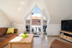 A modern loft apartment with a sloped ceiling, a wooden coffee table with a potted plant, a brown leather sofa, a television on a wooden stand, and a view of a tiled roof through a glass door.
