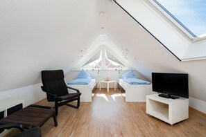 A modern attic bedroom with a skylight, a black chair, a bed with blue bedding, a small table, and a TV on a white stand.