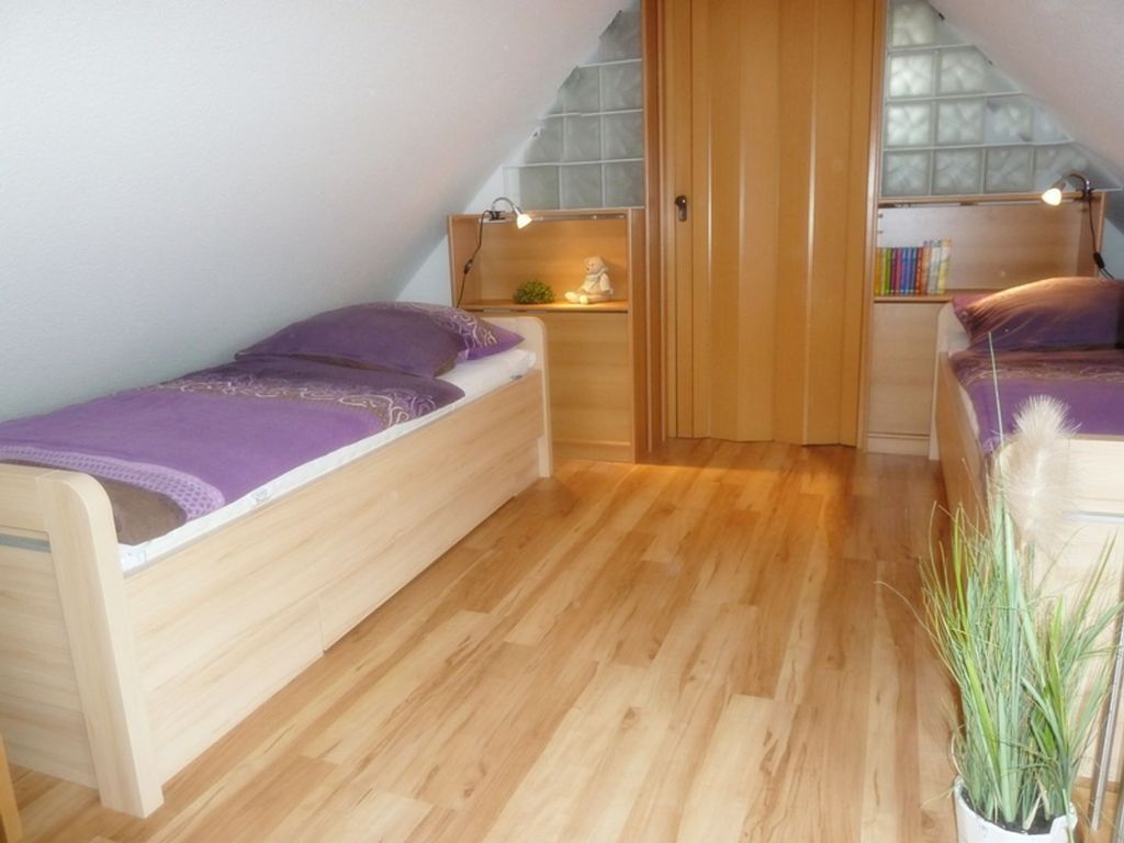 A small, cozy bedroom with two wooden beds, a wooden wardrobe, and a small shelf with books and a lamp.
