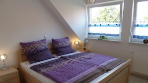 Un dormitorio con cama de madera, ropa de cama morada, una mesita de noche con lámpara y una ventana con vistas a zonas verdes.