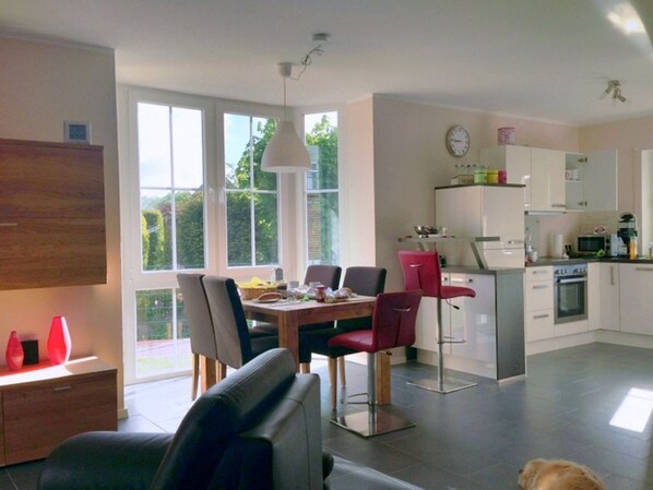 A modern kitchen with white cabinets, a dining area with a wooden table and chairs, and a large window providing a view of greenery outside.