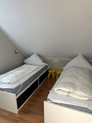 A small, compact room with two single beds, a wooden floor, and a small yellow stool.