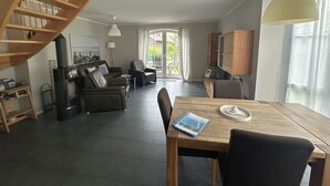 En rummelig opholdsstue med en trætrappe, et spisebord med en bog og udsigt gennem en glasdør til en udendørs terrasse.
