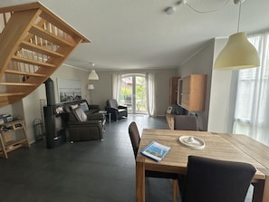 A spacious living area with a wooden staircase, a dining table with a book, and a view through a glass door to an outdoor patio. - Sieloase - End terraced house in a quiet location with water access (Krummhörn)
