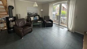 En stue med sofa, lænestole, et sofabord i glas, en pejs og udsigt gennem en skydedør til en udendørs terrasse.