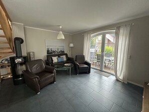 A living room with a sofa, armchairs, a glass coffee table, a fireplace, and a view through a sliding glass door to an outdoor patio. - Sieloase - End terraced house in a quiet location with water access (Krummhörn)
