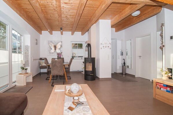 Una sala de estar moderna con techo de madera, comedor con mesa y sillas, estufa de leña negra y vistas amplias a través de grandes ventanales.