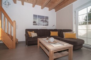 A modern living room with a wooden staircase, a brown sofa set, a wooden coffee table, and a large window with blinds. - Ferijentied - Kinderfreundliches Ferienhaus in Zentraler Lage (Krummhörn)