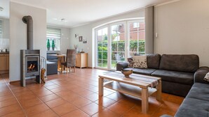 A spacious living room with a fireplace, a sofa, a dining area, and a large window offering a view of the outdoors. - Haus Otter - Beautiful vacation home (100sqm) for 6 persons and dog with fireplace, sauna (Norden)