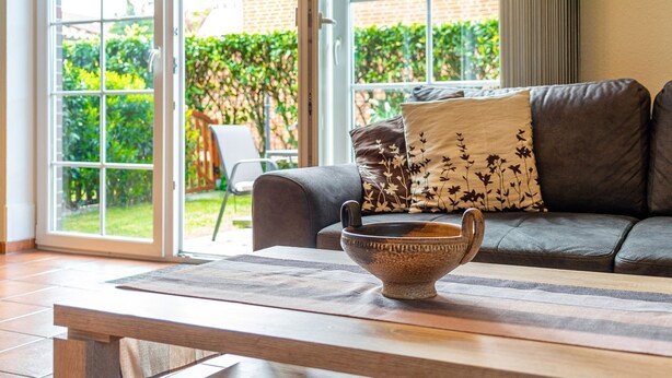 A living room with a sofa, a coffee table, and a view of a garden through large windows. - Haus Otter - Beautiful vacation home (100sqm) for 6 persons and dog with fireplace, sauna (Norden)