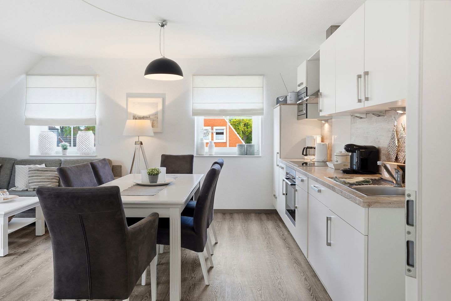 Eine moderne Küche mit weißen Schränken, ein Essbereich mit weißem Tisch und Stühlen und Blick auf ein Ziegelgebäude durch ein Fenster.
