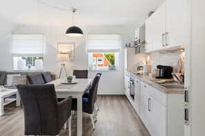A modern kitchen with white cabinets, a dining area with a white table and chairs, and a view of a brick building through a window.