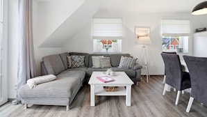 A modern loft-style living room with a gray sectional sofa, white coffee table, and a dining area with a white table and black chairs.