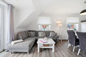 A modern loft-style living room with a gray sectional sofa, white coffee table, and a dining area with a white table and black chairs.