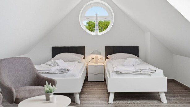 A small, modern bedroom with two single beds, a nightstand, a lamp, a chair, and a view through a circular window.