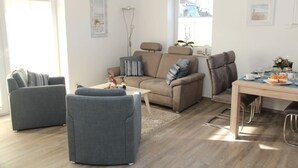 Een woonkamer met een bank, twee fauteuils, een salontafel en een eettafel met stoelen.