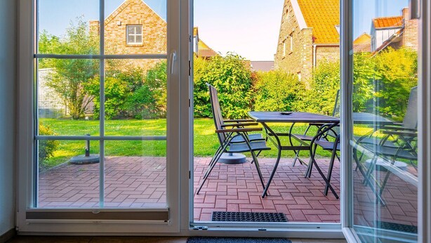 Ein Wintergarten mit Glastür, einem Patio-Tisch und -Stühlen, mit Blick auf eine Ziegelterrasse und einen Garten mit einem Ziegelhaus im Hintergrund.