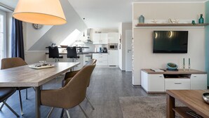 A modern kitchen with white cabinets, a dining area with a wooden table and chairs, a TV mounted on the wall, and a small shelf with decorative items.
