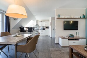 Una cocina moderna con armarios blancos, un comedor con mesa y sillas de madera, un televisor montado en la pared y un pequeño estante con adornos.