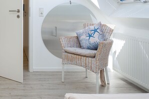 Una silla de mimbre con un cojín y un almohada estampada con motivos de estrellas, una puerta blanca y una pared blanca con un ventanal circular.