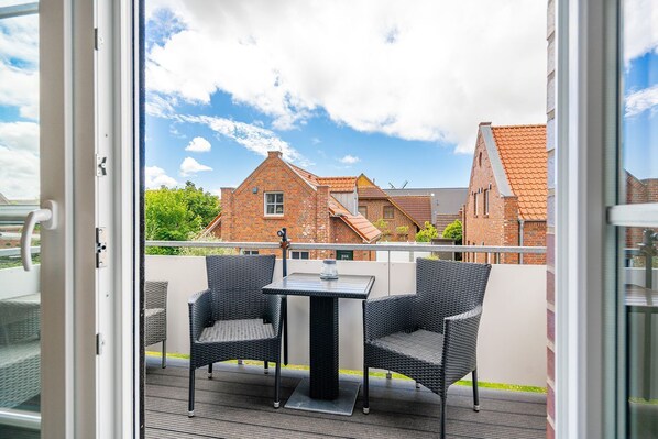 A balcony with wicker chairs and a table, overlooking a row of brick houses with red roofs.