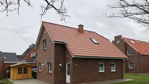 A two-story brick house with a red-tiled roof, a small wooden structure to the left, and a paved driveway. - Fiete - Beautiful vacation home directly in the village center! (Krummhörn)