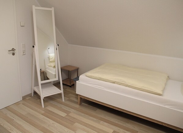 A bedroom with a single bed, a wooden floor, a white door, and a small wooden nightstand.
