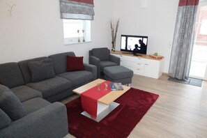 A modern living room with a grey sofa, a red ottoman, and a wooden coffee table. There is a flat-screen TV on a white cabinet, and a window with a view of a building. - Fiete - Beautiful vacation home directly in the village center! (Krummhörn)