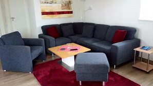 A modern living room with a grey sectional sofa, a small wooden coffee table, a grey ottoman, a red rug, and a painting on the wall. - Fiete - Beautiful vacation home directly in the village center! (Krummhörn)