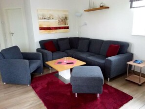 A modern living room with a grey sectional sofa, a small wooden coffee table, a grey ottoman, a red rug, and a painting on the wall. - Fiete - Beautiful vacation home directly in the village center! (Krummhörn)