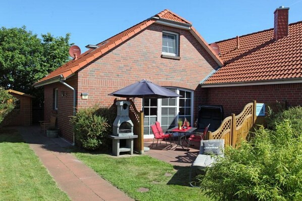 A brick house with a red-tiled roof, a barbecue grill, and a patio with red chairs and a table.