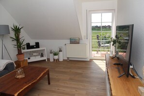 A modern living room with a wooden floor, white walls, a large window, and a view of a green landscape. - Deichblick 2 - High-quality vacation apartment with a wonderful view of the countryside (Krummhörn)