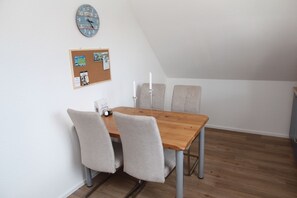 A dining area with a wooden table and four chairs, a corkboard with photos and notes, and a clock on the wall.
