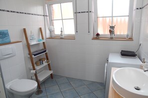 A bathroom with a toilet, sink, and shelves with toiletries. There are windows with curtains and a view of trees outside.