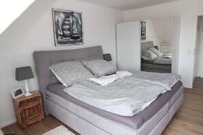 A bedroom with a bed, a nightstand, a lamp, a painting, and a wardrobe. - Deichblick 2 - High-quality vacation apartment with a wonderful view of the countryside (Krummhörn)