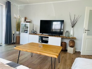 Un salon moderne avec une table basse en bois, un téléviseur à écran plat fixé sur une étagère en bois, des meubles blancs et une grande fenêtre avec des rideaux.