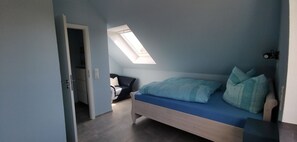 Une chambre avec un puits de lumière, un lit avec une literie bleue, une chaise et une porte donnant sur une autre pièce.