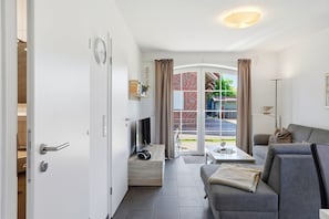Ein modernes Wohnzimmer mit einer Couch, einem Fernseher und einem Esstisch. Eine Schiebetür aus Glas führt zu einem Außenbereich mit Blick auf ein Backsteinhaus und viel Grün.
