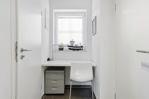 A narrow room with a white desk, a chair, a window with blinds, and a small shelf with a model ship.