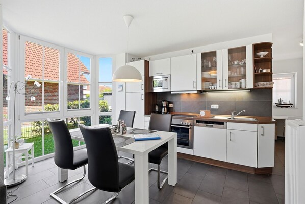 Eine moderne Küche mit weißen Schränken, ein Essbereich mit Tisch und Stühlen sowie ein Blick ins Freie durch große Fenster.
