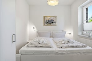 Un lit bien fait, avec du linge de lit et des serviettes blanches, une table de chevet, un luminaire fixé au mur et une fenêtre avec des stores.