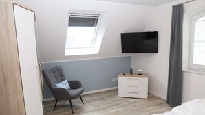 Een slaapkamer met een bed, een stoel, een nachtkastje, een televisie en een raam met jaloezieën.