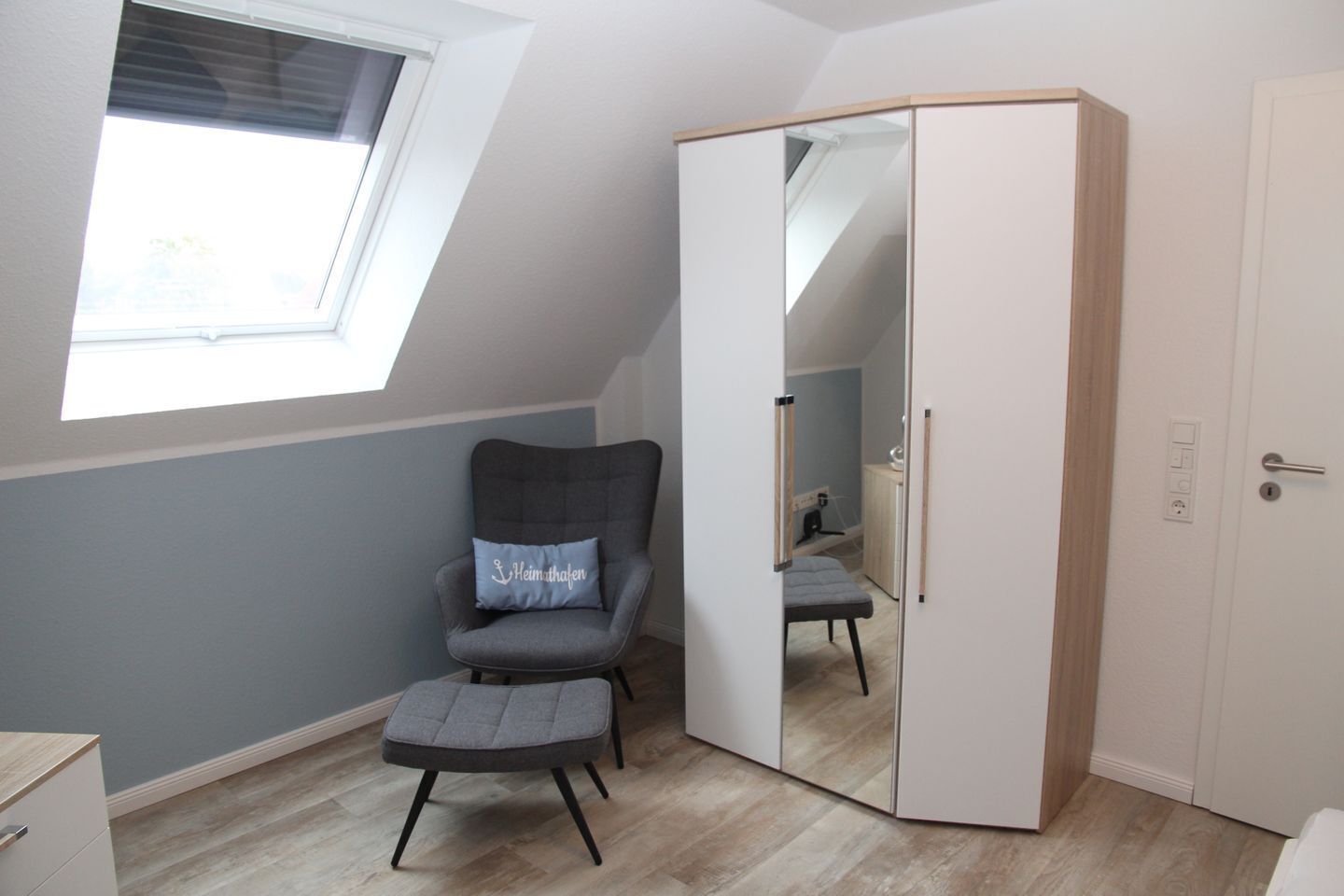 A room with a sloped ceiling, a window, a grey armchair with a cushion, a small stool, and a wardrobe with a mirror.