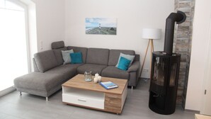 Een moderne woonkamer met een grijze hoekbank, een houten salontafel, een zwarte houtkachel en een schilderij aan de muur.