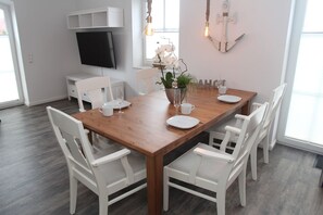 Un comedor con una mesa de madera, sillas blancas, un televisor montado en la pared y una ventana que deja entrar luz natural.